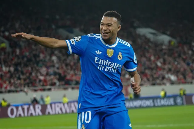 Un fotbalist într-un echipament sportiv albastru cu logo-uri de sponsor și un scut pe piept zâmbește cu brațele întinse pe un teren de fotbal.