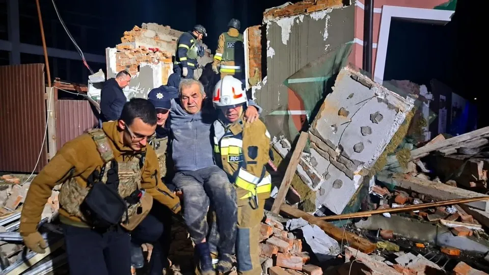 Trei bărbați, dintre care unul poartă echipament de protecție, susțin un bărbat în haine gri printre dărâmături de cărămizi și resturi de construcții noaptea.