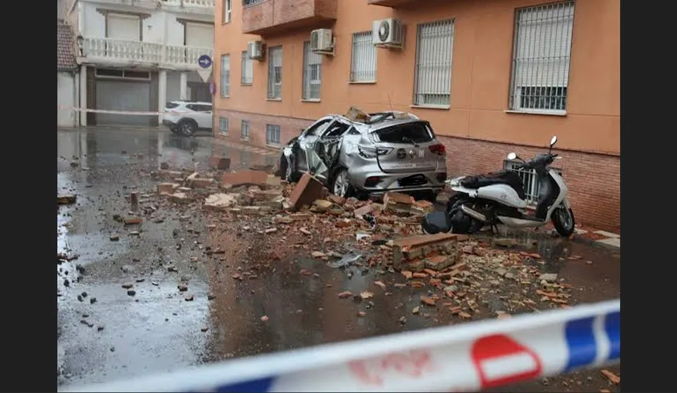 Imaginea arată o mașină avariată și un scooter parcate lângă un clădiră cu dărâmături pe carosabil, în spatele unei benzi de poliție.