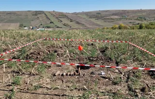 Un teren agricol cu resturi vegetale este delimitat de o bandă de alertă albă și roșie, având obiecte metalice cilindrice pe sol.