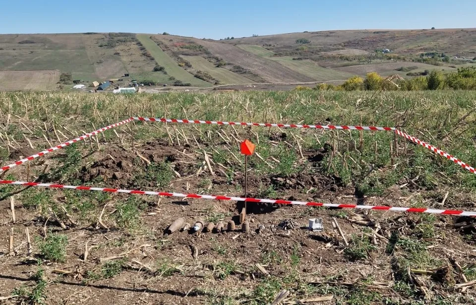 Un teren agricol cu resturi vegetale este delimitat de o bandă de alertă albă și roșie, având obiecte metalice cilindrice pe sol.