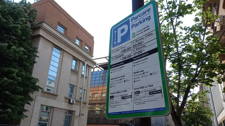 Un panou informativ de parcare verde este montat pe un stâlp lângă o clădire și un copac.