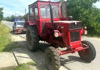 Un tractor roșu cu cabină de sticlă este parcat pe marginea unui drum lângă un vehicul alb.