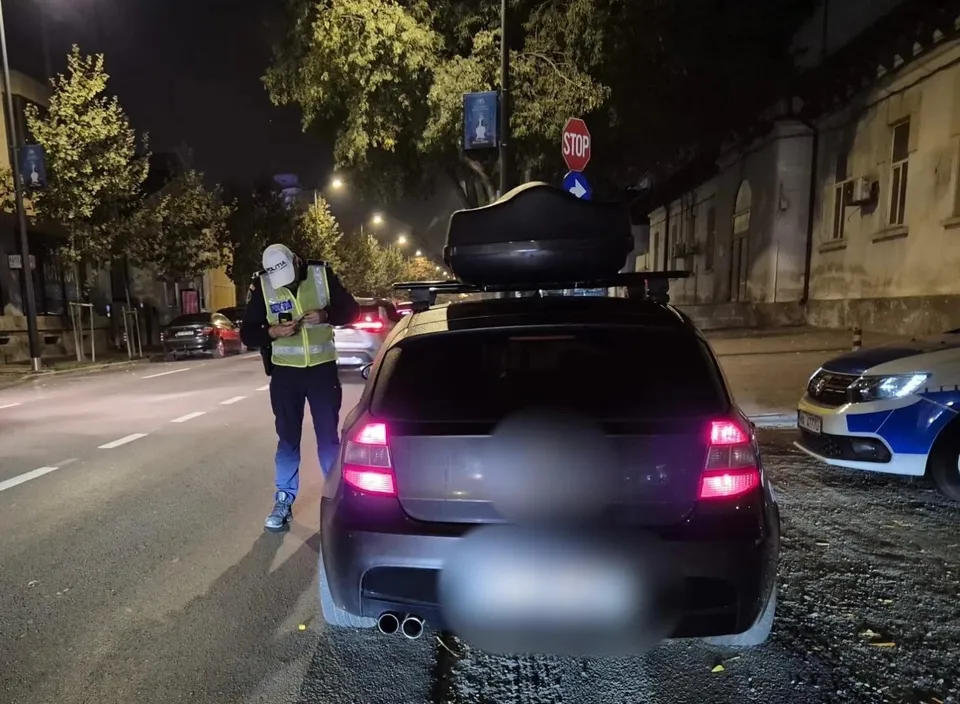 Un agent de poliție cu vestă reflectorizantă stă lângă un autovehicul negru cu cutie de transport pe plafon, pe un drum urban noaptea.