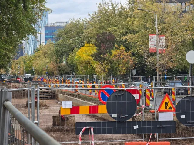 Un șantier este delimitat de garduri metalice, conuri de trafic și semne de circulație pe un drum cu mașini în fundal.