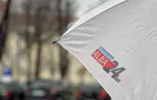 O umbrelă albă cu un logo colorat este poziționată în prim-plan, având un fundal urban desfocat.