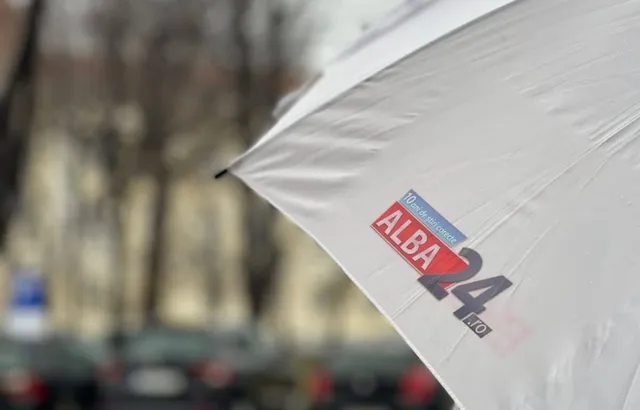 O umbrelă albă cu un logo colorat este poziționată în prim-plan, având un fundal urban desfocat.