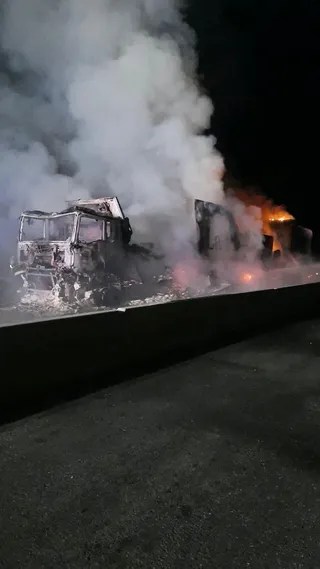 Un autocamion complet ars, cu cabina distrusă, emiteând un volum mare de fum alb și gri într-un mediu noapte, văzut din lateral peste un bord de beton.