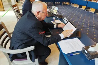Un bărbat cu ochelari și costum închis stă la o masă albastră, citind un document de pe o tabletă.