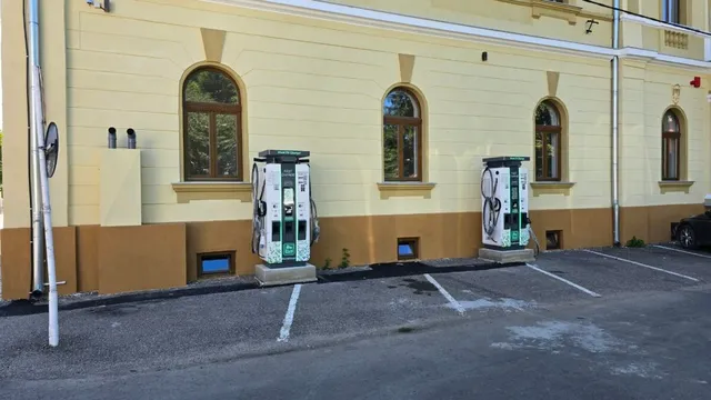 O clădire galbenă cu ferestre arcuite prezintă două stații de încărcare pentru vehicule electrice amplasate pe un parcare asfaltată în fața acesteia.
