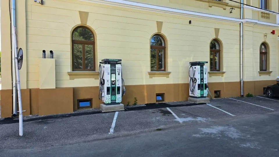 O clădire galbenă cu ferestre arcuite prezintă două stații de încărcare pentru vehicule electrice amplasate pe un parcare asfaltată în fața acesteia.