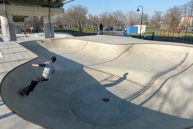 Un bărbat cu tricou alb și pantaloni închiși se află pe o rampă de beton într-un skatepark sub un viaduct.