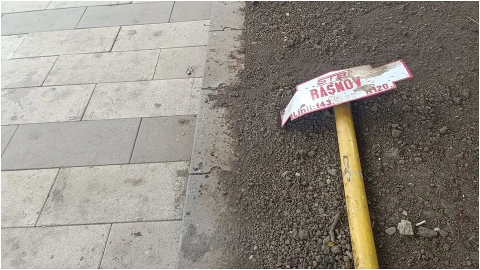 Un semn alb cu text roșu este sprijinit pe un stâlp galben în interiorul unui spațiu cu pământ lângă un trotuar pavat.