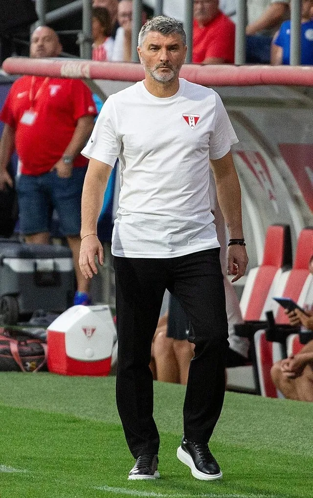 Un bărbat cu părul scurt și barbă căruia îi este îmbrăcat un tricou alb și pantaloni negri merge pe un teren de fotbal.