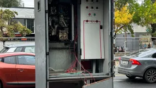 Un dulap electric metalic deschis prezintă cabluri colorate expuse și componente interne vizibile pe un fundal stradal cu mașini și copaci.