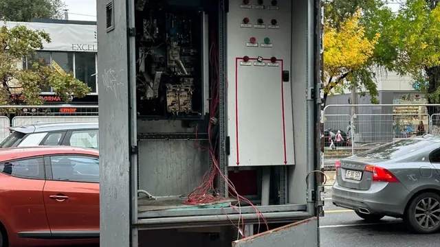 Un dulap electric metalic deschis prezintă cabluri colorate expuse și componente interne vizibile pe un fundal stradal cu mașini și copaci.