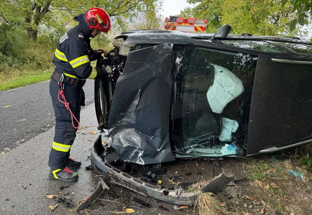 Un salvator cu cască roșie și costum de lucru în culori închise stă lângă un autoturism gri răsturnat pe o porțiune de drum asfaltat.