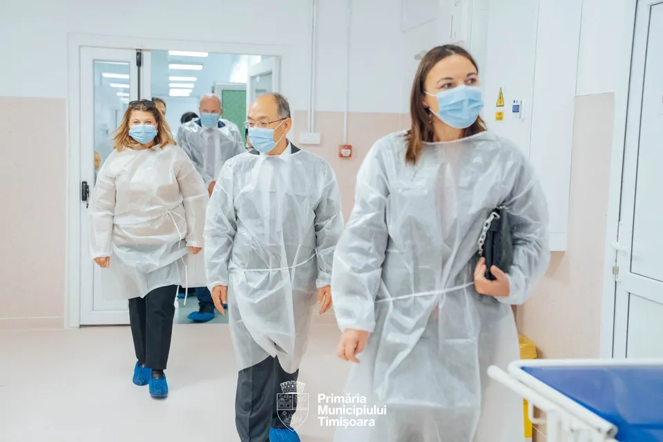 Patru persoane purtând halate medicale de protecție și măști faciale parcurg un hol cu pereți albi.