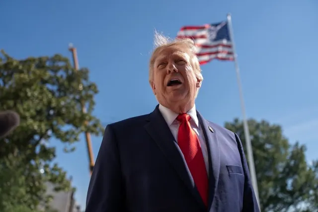 Un bărbat cu părul blond, purtând costum închis la culoare și cravată roșie, stă cu gura deschisă sub un cer senin cu un steag american.