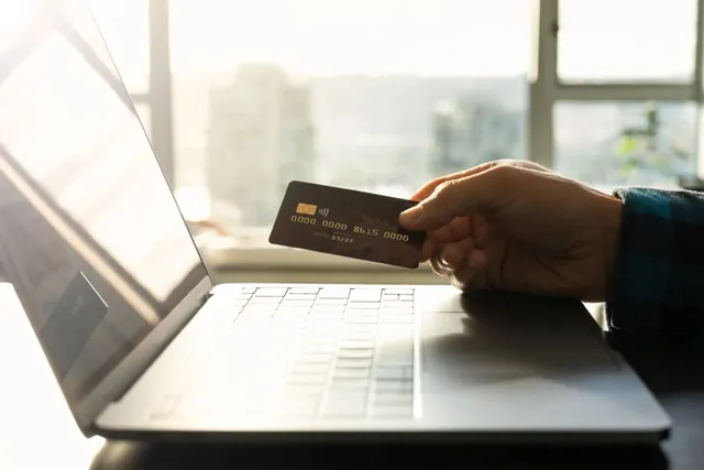 O mână ține un card bancar deasupra unui laptop deschis într-o încăpere luminoasă.