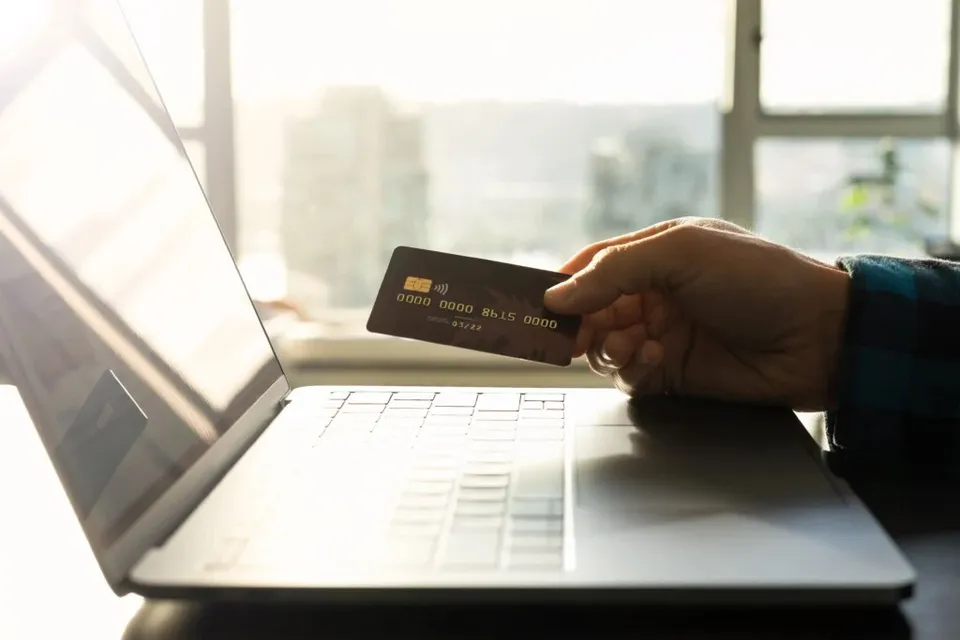 O mână ține un card bancar deasupra unui laptop deschis într-o încăpere luminoasă.