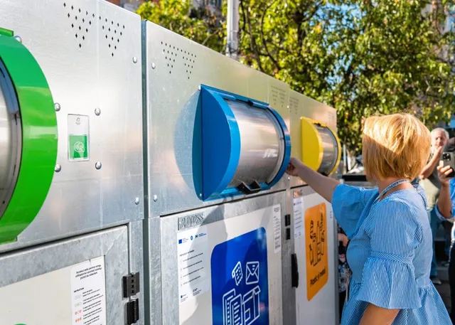 O femeie cu părul scurt și bluză albastră deschide un recipient de reciclare albastru de pe un ansamblu de containere metalice de colectare.