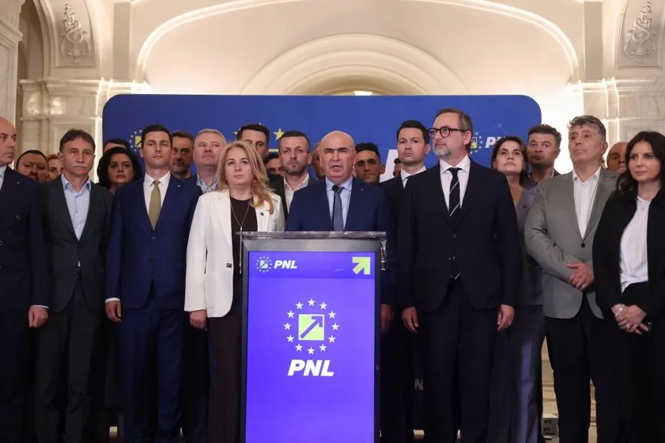 Un grup de oameni în haine formale stă în fața unui panou albastru cu logo-ul PNL și a unui pupitru cu același simbol.
