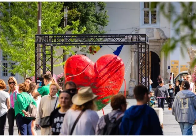 O structură metalică susține un obiect roșu în formă de inimă în mijlocul unei mulțimi de oameni într-un spațiu public exterior.