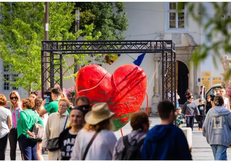 O structură metalică susține un obiect roșu în formă de inimă în mijlocul unei mulțimi de oameni într-un spațiu public exterior.