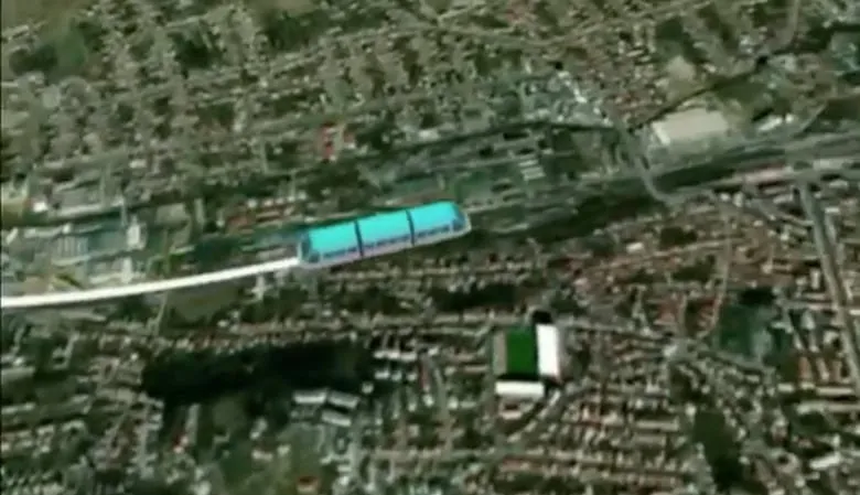 O vedere aeriană deasupra unui oraș prezintă o structură lungă și subțire de culoare albă cu secțiuni albastre, situată deasupra clădirilor și a vegetației.