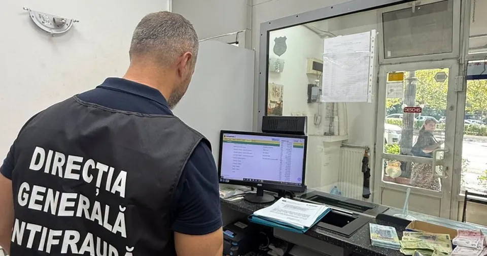 Un bărbat cu vestă neagră pe care scrie "DIRECȚIA GENERALĂ ANTIFRAUDĂ" stă cu spatele la cameră, privind un monitor de calculator, într-un spațiu ce pare a fi o casă de schimb valutar, cu bani pe tejghea.