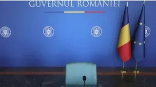 Un scaun de culoare deschisă este poziționat în fața unui perete albastru pe care sunt afișate sigilii și textul Guvernului României, lângă două steaguri.