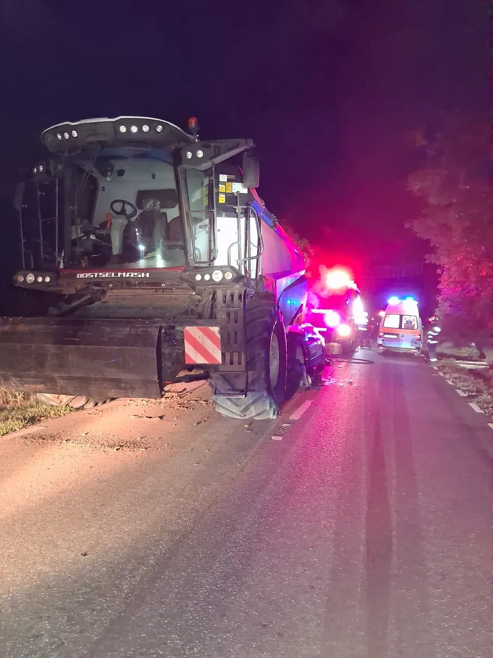 O combină agricolă este oprită pe marginea unui drum lângă un autoturism, în timp ce vehicule de intervenție cu lumini colorate sunt vizibile în fundal.