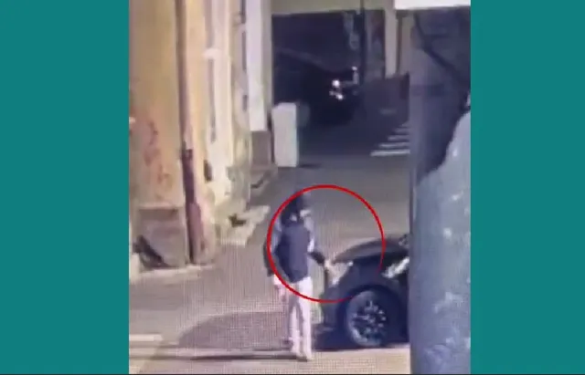 O persoană cu haine închise stă lângă un autoturism negru pe un trotuar lângă un zid de clădire sub lumină nocturnă.