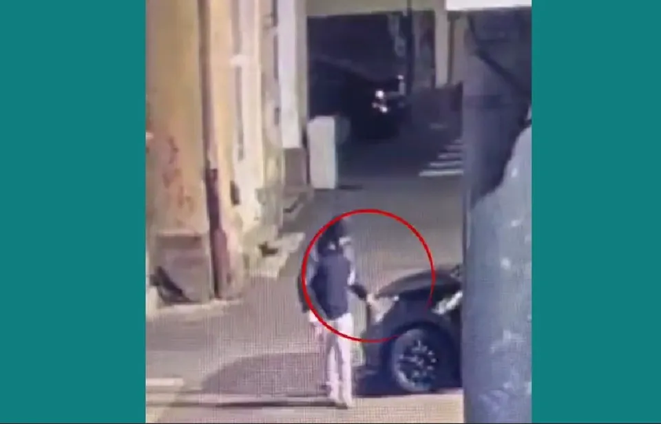 O persoană cu haine închise stă lângă un autoturism negru pe un trotuar lângă un zid de clădire sub lumină nocturnă.