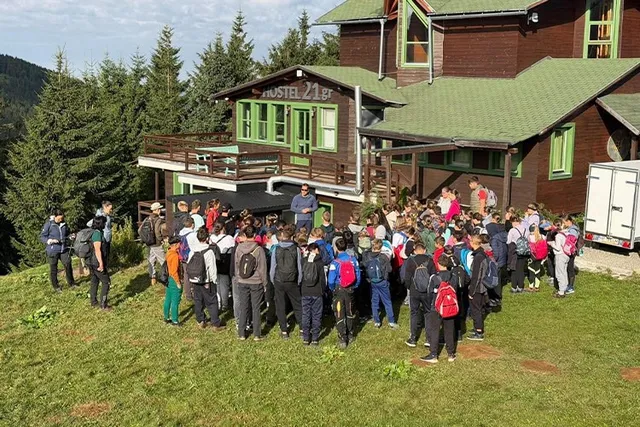 Un grup numeros de persoane stă adunat pe o iarbă verde în fața unei clădiri de lemn cu acoperiș verde, înconjurat de pădure.