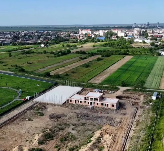 O vedere aeriană prezintă un teren de construcție cu o clădire de cărămidă și o structură acoperită cu un panou alb lângă câmpuri verzi.
