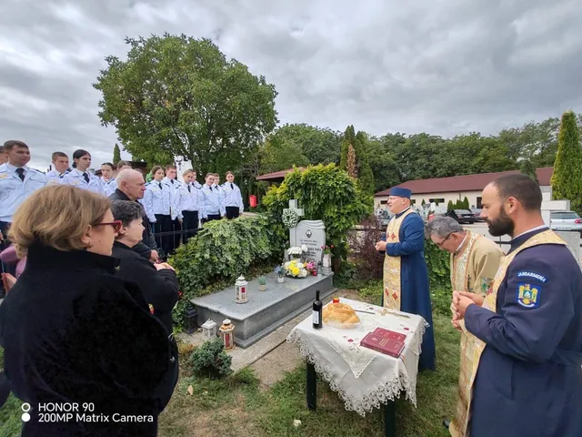 Un grup de persoane stă în fața unei morminte, în timp ce doi bărbați în haine religioase stau lângă o masă cu obiecte ritualice.