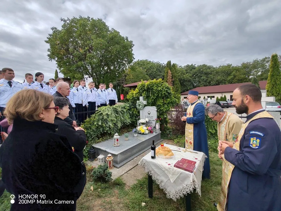 Un grup de persoane stă în fața unei morminte, în timp ce doi bărbați în haine religioase stau lângă o masă cu obiecte ritualice.