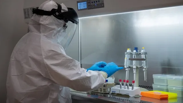 O persoană îmbrăcată într-un costum alb de protecție cu vizieră manipulează recipiente mici în interiorul unei cabine de laborator cu lumină albă.