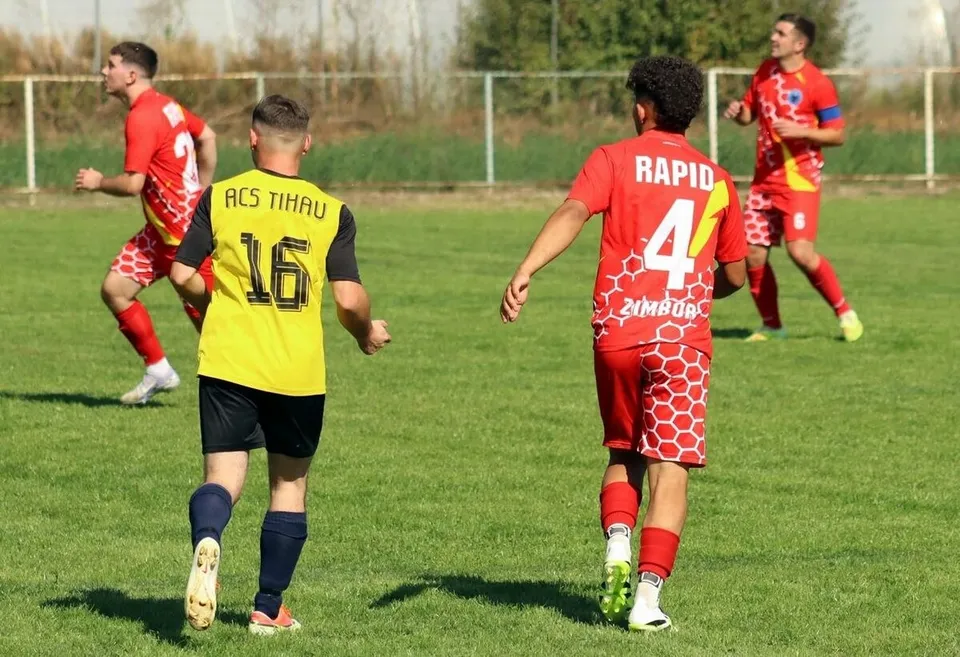 Patru jucători de fotbal cu echipamente roșii și galben pe un teren de iarbă verde sunt dispersați în câmpul de joc.