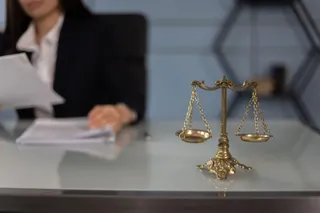 O balanță de metal este așezată pe o masă de birou lângă un dosar, în timp ce o persoană în costum stă în fundal.