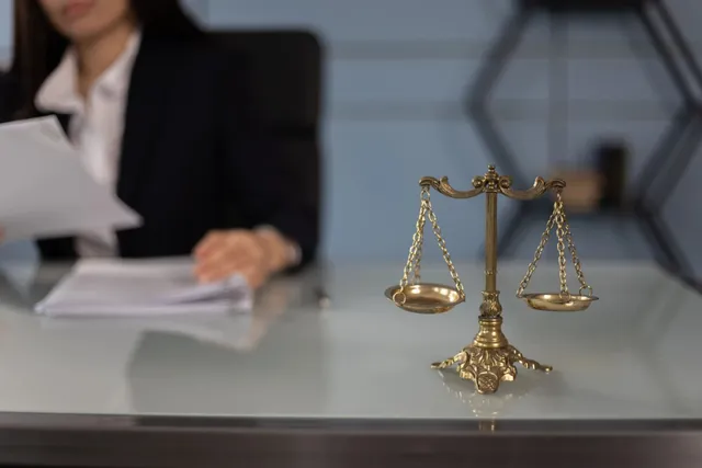 O balanță de metal este așezată pe o masă de birou lângă un dosar, în timp ce o persoană în costum stă în fundal.