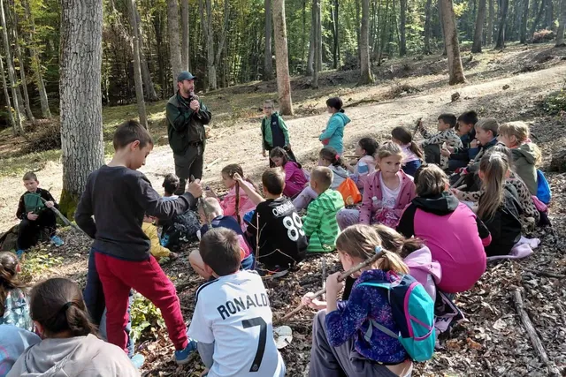 Un grup de copii stă așezat pe un teren împădurit, privind spre un bărbat în picioare care poartă o vestă neagră.