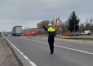 Un polițist cu vestă reflectorizantă stă pe marginea unei drumuri asfaltate lângă un camion și un autoturism sub un cer înnorat.