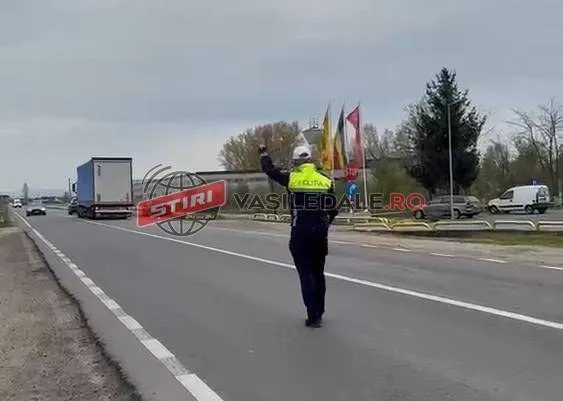 Un polițist cu vestă reflectorizantă stă pe marginea unei drumuri asfaltate lângă un camion și un autoturism sub un cer înnorat.