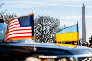 Un steag american și un steag ucrainean sunt montate pe capota unei mașini negre, având un obelisc și copaci în fundal.