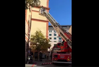 Un autospecial de pompieri cu o scară extensibilă ridicată este poziționat lângă o clădire înaltă, în prezența unor persoane în uniformă.