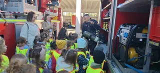 Un grup de copii poartă veste fluorescente și ascultă un bărbat în uniformă întunecată lângă un vehicul de serviciu cu echipamente de salvare.