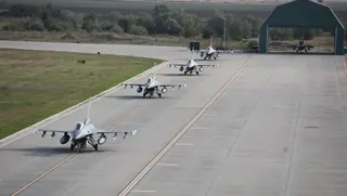 Trei avioane de vânătoare gri taxiază pe o pistă de beton lângă o zonă cu iarbă și un hangar deschis.